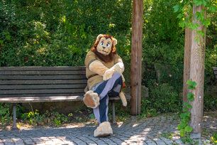 Wimasimba sitzt als Löwe im Fursuit auf einer Parkbank im Schatten.